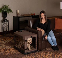 a happy woman holding the gate of the chocolate colored steel dog crate open sitting on the leather couch in a modern living room with bookshelf and a flowerpot at the back