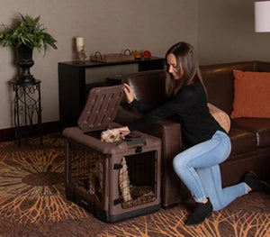 a woman kneeling on the floor holding the top cover of a chocolate colored steel dog crate open with her dog inside next to a leather couch in a modern living room with book shelf and a flower pot at the back