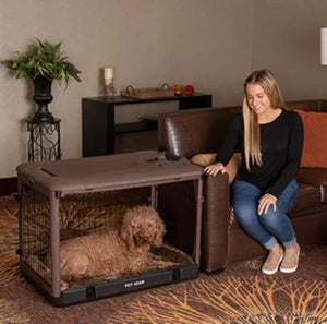 a happy woman sitting on a leather couch staring at her dog laying inside a chocolate colored steel dog crate and a flower pot on the background