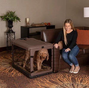 a happy woman sitting on a leather couch next to her dog inside a chocolate colored steel dog crate holding the steel gate of the crate open for her dog in a modern living room setting
