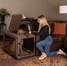 a happy woman playing with her dog inside a chocolate  colored steel dog crate next to a leather couch and a pot of flower on the background