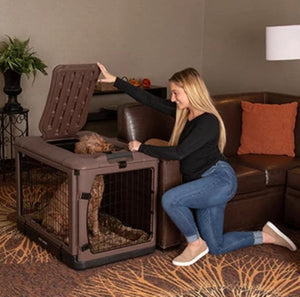 a happy woman playing with her dog inside a chocolate  colored steel dog crate next to a leather couch and a pot of flower on the background