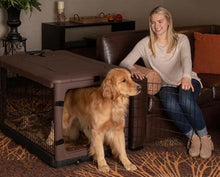 a happy woman sitting on a leather couch holding the steel door open for her dog to get out of a  large brown dog steel crate in a modern living room with bookshelf at the back and flower pot 