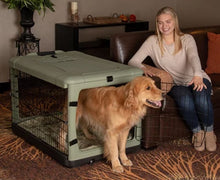 a happy woman sitting on a leather couch holding the steel door open for her dog to get out of a large brown dog steel crate in a modern living room with bookshelf at the back and flower pot