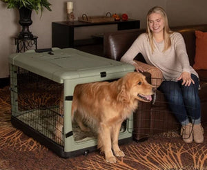 a happy woman sitting on a leather couch next to her dog inside a sage colored steel dog crate holding the steel gate of the crate open for her dog in a modern living room setting