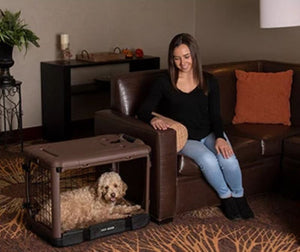 a woman siting on a leather couch staring ate her dog inside a chocolate colored steel dog crate with bolster pad in a modern living room with wooden shelf and a pot of flower at the back