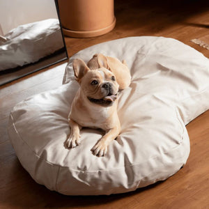 a french bulldog laying on a cream colored dog bed on a wooden floor next to a wooden couch