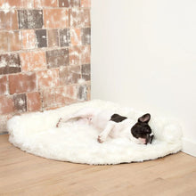 a french bulldog laying on a white furry dog bed on the corner of a white and brick wall