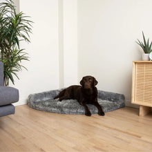 a black labrador siting on grey furry dog bed on the corner of the house in between of a couch and a wooden drawer with plants on them