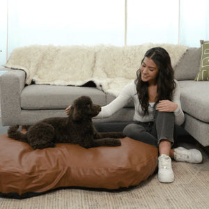 a woamn sitting next to her dog laying on a brown leather dog bed next to a grey couch with a white dog blanket on it in an all white modern room 