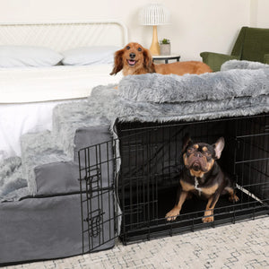 a fluffy dachshund sitting on top of a grey fluffy bedside sleeper and a french bulldog sitting inside a balck dog steel crate next to a white bed 