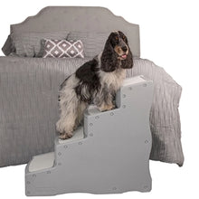 a fluffy dog standing on a four step grey dog stair next to a grey bed with white background