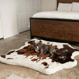 a french bulldog laying on a fluffy dog bed with cowhide print next to a wooden bed in a white bedroom
