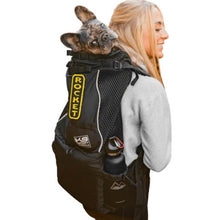 a lady in white carrying her dog on her back in a black dog backpack carrier with water bottle on it's side pocket