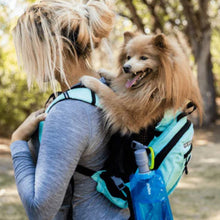 a lady carrying her dog on her back in a blue dog backpack carrier with a portable dog water bottle on the side pockets walking in the woods 