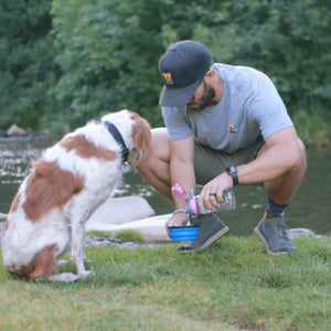 a man putting some water on a blue dog saucer bowl next to his dog and the river