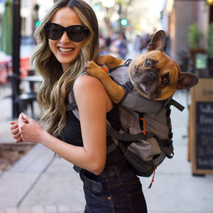 A happy woman wearing sunglasses with her dog in a grey dog backpack carrier in her back walking on a sidewalk