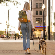 A woman walking her dog on a leash carrying a leafy green backpack on the sidewalk