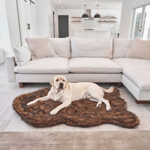 a white labrador retriever laying on a brown furry dog bed next to an L shaped couch in an all white modern living room
