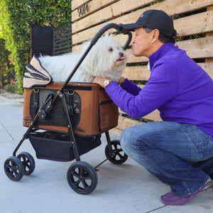 a man kissing his dog inside a brown dog carrier/stroller next to a wooden gate 