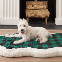 A white terrier in front of a fireplace laying on a white furry dog bed with green and black checkered pattern