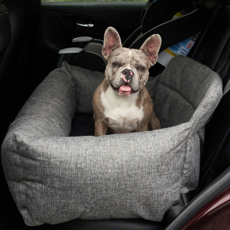 A french bulldog sticking his tongue out sitting on a grey dog bed in the car seat