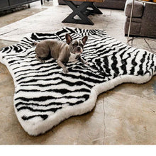 A French bulldog laying on a Zebra  printed furry curved dog bed in a living room next to a wooden table and black cabinets