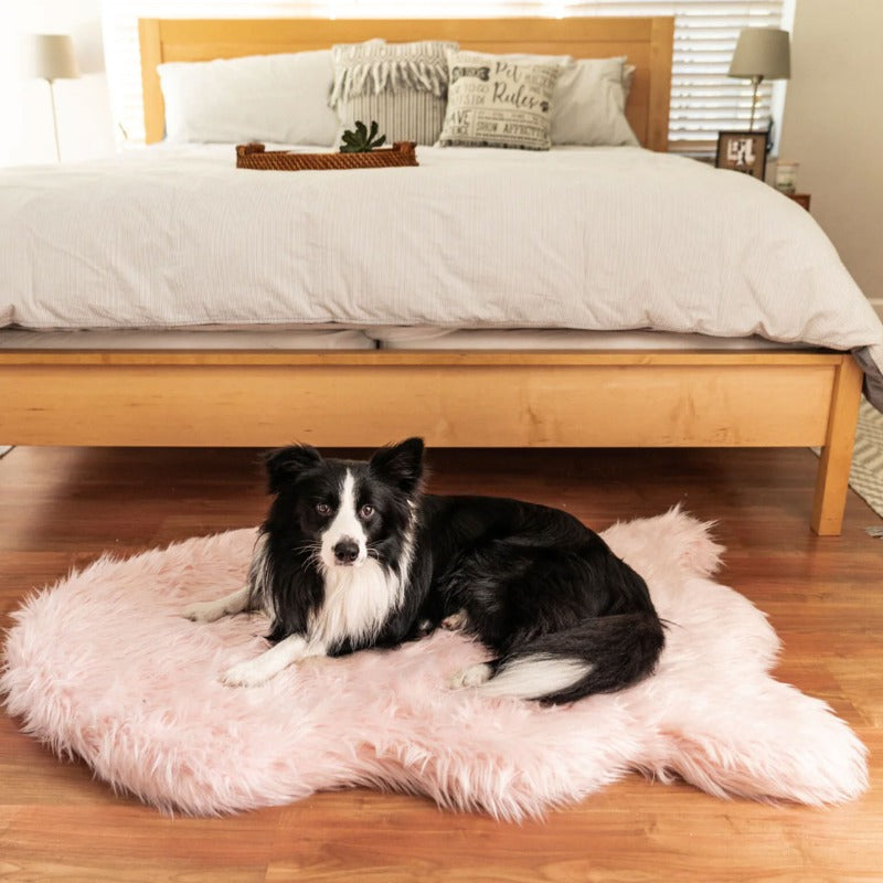 A border collie on the floor of a modern bedroom laying on a furry curved Blush pink dog bed in front of a wooden bed with white foam and pillow and two lamps at the back