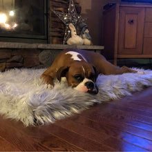 A boxer on the floor next to a fireplace and wooden drawers on a christmas setting laying on furry grey rectangular dog bed 