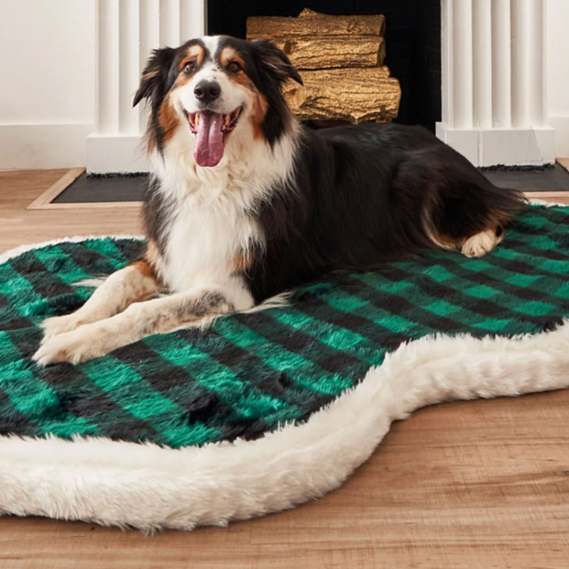 An australian shepperd in front of a fireplace laying on a white furry dog bed with green and black checkered pattern