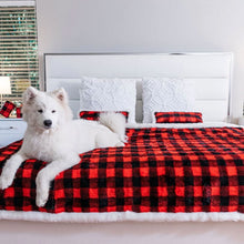 A white samoyed on the edge of a white bed on a bedroom laying on a red and black checkered pattern waterproof dog blanket with a tiny giftbox and a lamp on the background near the window 