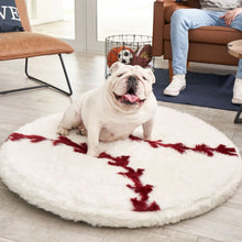 a white English bulldog in a living room sitting on a round baseball shaped furry dog bed next to a brown chair, sofa  and a bin of balls and a sitting man with white shoes and blue jeans 