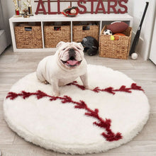 A white english bulldog sitting at the edge of a round baseball shaped dog bed with a background of baseball items , trophies and all star text on a white four dividers and a basket full of balls and a black baseball helmet