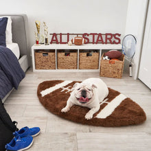 A white english bulldog in a room next to a blue pair of shoe and a bed laying in front of a drawer with a basket full of toys and a tennis racket on a football shaped dog bed 