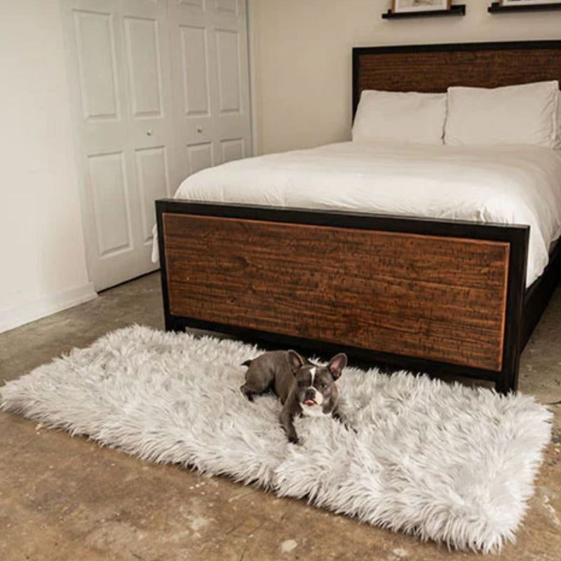 A french bulldog in a bedroom laying on a rectagular grey dogbed next to a wooden bed with white foam and pillows on it 
