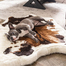 A French bulldog on the floor laying on a soft furry white dog bed with dark brown and tan cow hide pattern next to a black triangular frame 