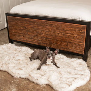 A french bulldog laying on a furry white with brown accent dog bed next to a wooden bed with white foam next to it on a bedroom