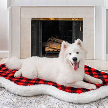 A white Samoyed in front of a fireplace laying on a furry dog bed with red and black checkered pattern