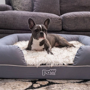 A French Bulldog laying on gray rectagular dog bed with white with brown accent fur matt in front of a grey couch