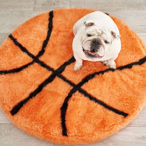 A top view of a english white bulldog sticking his tongue out sitting on a basketball shaped dog bed