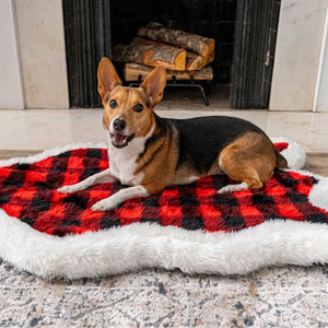 A brown corgi with black back fur in front of a fireplace laying on a furry dog bed with red and black checkered pattern