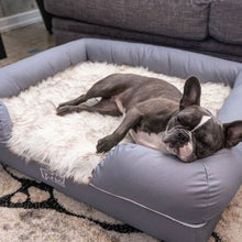 A French Bulldog sleeping on a rectagular dog bed with a white brown accents furry matt right next to a gray couch on a modern setting