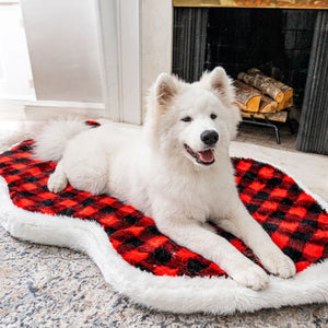 A white samoyed in front of a fireplace laying on a furry dog bed with red and black checkered pattern