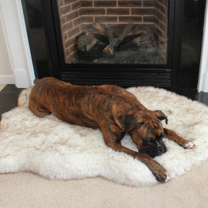 A boxer next to a fireplace laying on a furry white dog bed with brown accents