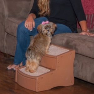 an image of a shih-tzu standing on a dog step next to a lady sitting on a grey couch 