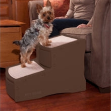 a yorkie standing on a dog steps next to a wooden drawer and a lady sitting on a grey couch