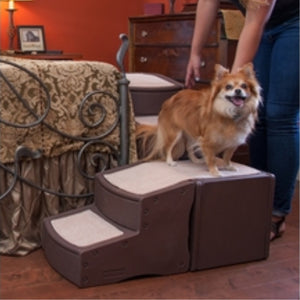 A Pomeranian on top of Pet Gear Easy Step Bed Stair, Chocolate next to a bed with grey steel frame and a lady next to wooden drawers 
