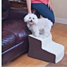 Close up image of a poodle on top of a dog stair Pet Gear Easy Step II Deluxe Soft Step, Oatmeal/Chocolate next to a brown leather couch and a leg of a lady in blue jeans 