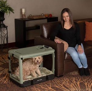 a woman siting on a leather couch staring ate her dog inside a sage colored steel dog crate with bolster pad in a modern living room with wooden shelf and a pot of flower at the back 