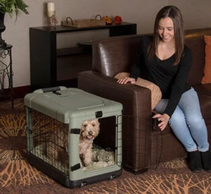 a happy woman holding the gate of the sage colored steel dog crate open sitting on the leather couch in a modern living room with bookshelf and a flowerpot at the back
